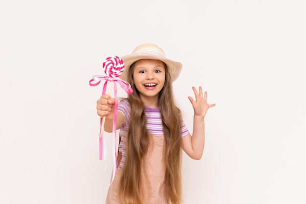 Una niña pequeña muestra una piruleta y sonríe Un niño con un vestido de verano y un sombrero de verano sobre un fondo blanco aislado