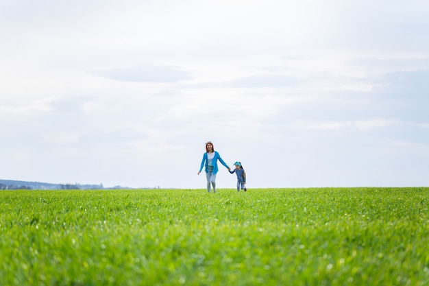 Niña pequeña y mujer madre corren y saltan, hierba verde en el campo, clima soleado de primavera, sonrisa y alegría del niño, cielo azul con nubes