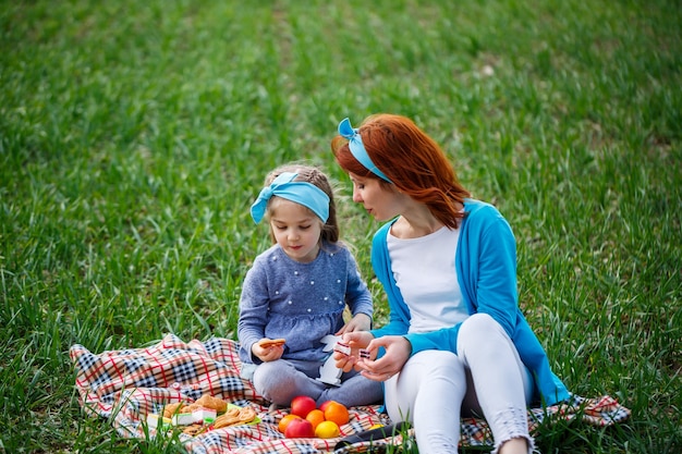 Niña pequeña y mujer madre sentada en la colcha y comiendo galletas y mermelada, pasto verde en el campo, clima soleado de primavera, sonrisa y alegría del niño, cielo azul con nubes