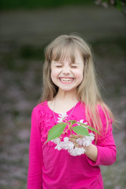 Niña pequeña o niño lindo con adorable rostro sonriente y cabello rubio en camisa rosa con flor de sakura de primavera flor de cerezo al aire libre en un fondo borroso