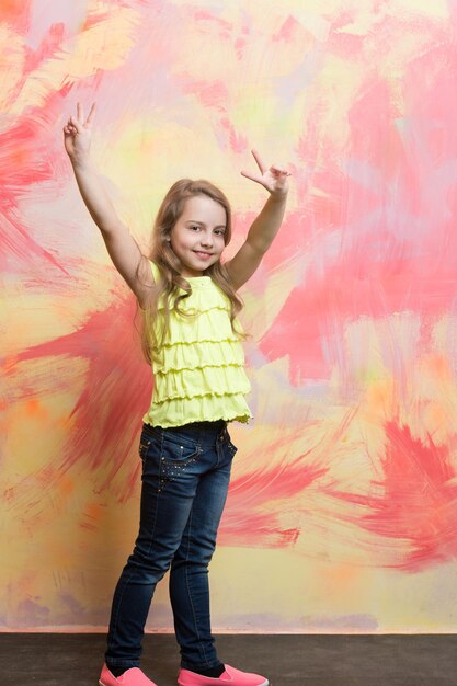 niña pequeña o niño lindo con cara feliz y cabello rubio en camisa amarilla y jeans sobre fondo abstracto colorido