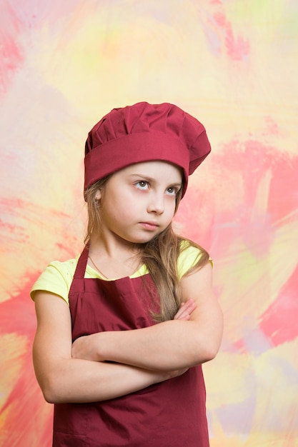 Niña pequeña o niño lindo con cara seria en gorro de cocinero rojo y delantal de cocinero sobre fondo abstracto colorido