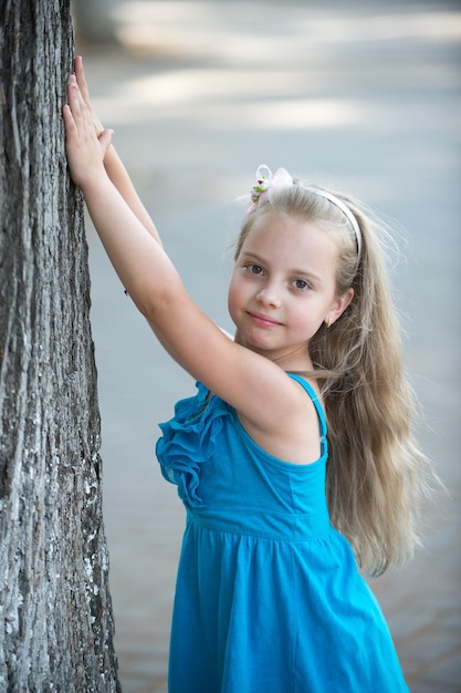Una niña pequeña o un niño lindo y feliz con una adorable cara sonriente y un arco en el pelo rubio con un vestido azul en verano al aire libre cerca de un árbol en un fondo borroso