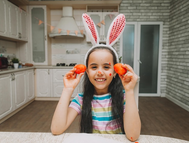 Una niña pequeña con orejas de conejo rosa sostiene huevos de pascua en la cara cerca de los ojos en la cocina