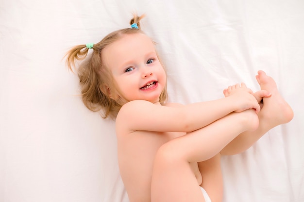 La niña pequeña en pañales sonríe, miente en cama en una sábana blanca