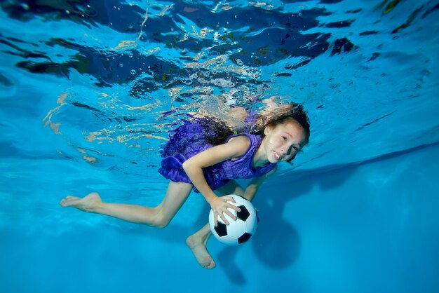 Una niña pequeña con una pelota de fútbol en las manos que practica deportes y nada bajo el agua en la piscina