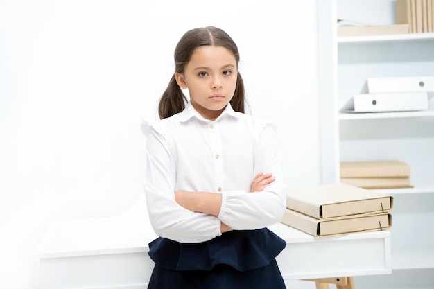 Niña pequeña pero seria viste uniforme escolar de pie con los brazos cruzados en el pecho Niña inteligente de colegiala se ve seria fondo interior blanco Niña seria sobre el conocimiento y la educación