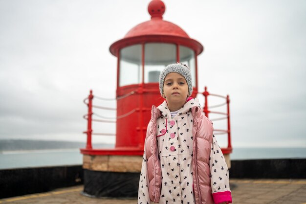 Niña pequeña de pie cerca del faro rojo en la orilla rocosa