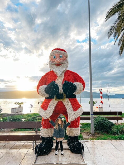 Foto niña pequeña de pie cerca de la figura de papá noel en el paseo marítimo por el mar