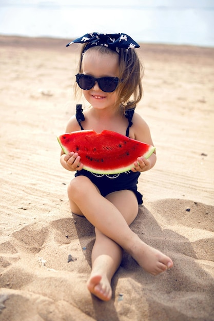 Foto niña pequeña de pie en la orilla de la playa gafas negras comiendo sandía jugosa