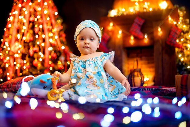 Niña pequeña en pijama sentada en cuadros escoceses cerca del árbol de Navidad y chimenea y guirnaldas