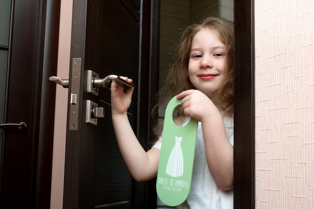 Niña pequeña con pintalabios pintado se asoma por detrás de la puerta que dice Vístete para impresionar No molestar