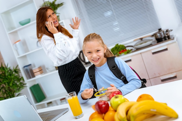 Una niña pequeña prepara un desayuno mientras su madre con exceso de trabajo llama por teléfono, antes de ir al trabajo. La madre se está enfadando por el estrés.