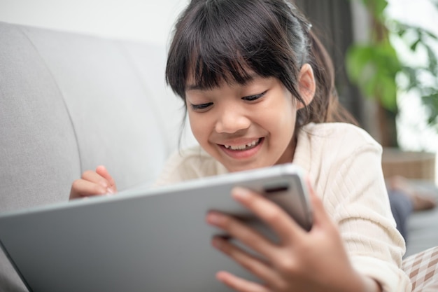 Niña pequeña que usa una tableta para jugar en Internet Niño sentado en el sofá viendo o hablando con un amigo en línea Niño relajándose en la sala de estar por la mañana Niños con el concepto de Nueva Tecnología