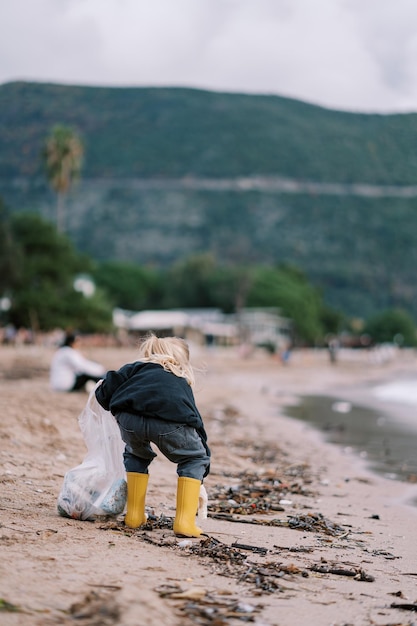 Niña pequeña recoge la basura en una bolsa mientras se agacha en la playa vista trasera