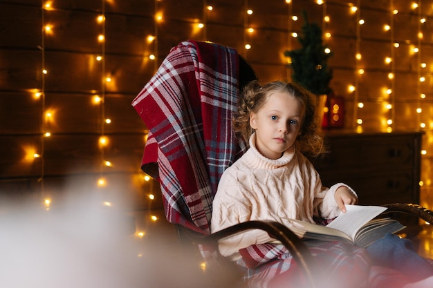 Niña pequeña rubia feliz y rizada envuelta en tela escocesa mirando a la cámara leyendo un libro sentado en una mecedora en una habitación oscura con un interior festivo