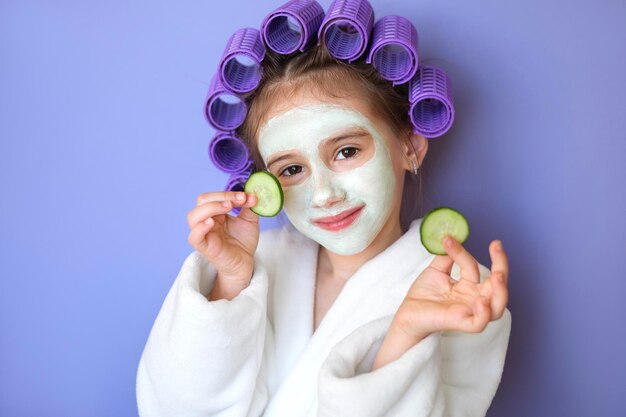 Niña pequeña con rulos y máscara facial de arcilla sostiene rodajas de pepino sobre fondo morado