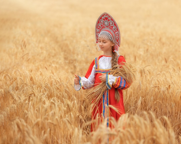 Niña pequeña en sarafan nacional ruso y un kokoshnik de pie en un campo de trigo dorado en el día de verano