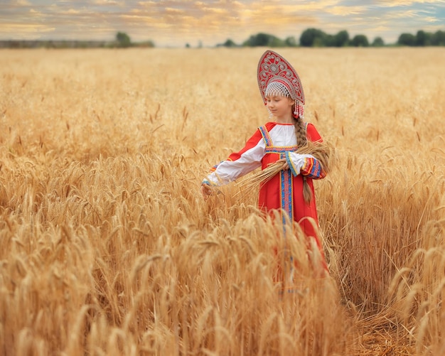 Niña pequeña en sarafan nacional ruso y un kokoshnik de pie en un campo de trigo dorado en summe