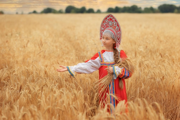 Niña pequeña en sarafan nacional ruso y un kokoshnik de pie en un campo de trigo dorado en summe