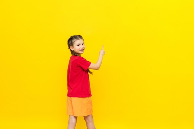 Una niña pequeña señala con el dedo un espacio vacío para su anuncio en un fondo amarillo aislado. Un hermoso niño sonríe en una camiseta roja con dos coletas. Copiar espacio.