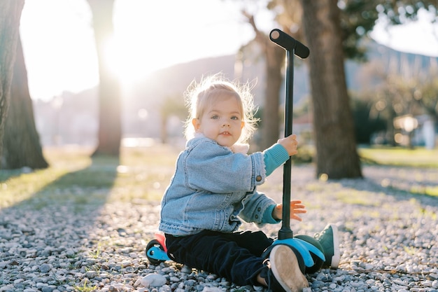 Niña pequeña sentada sosteniendo el volante de un scooter en un camino de guijarros en el parque