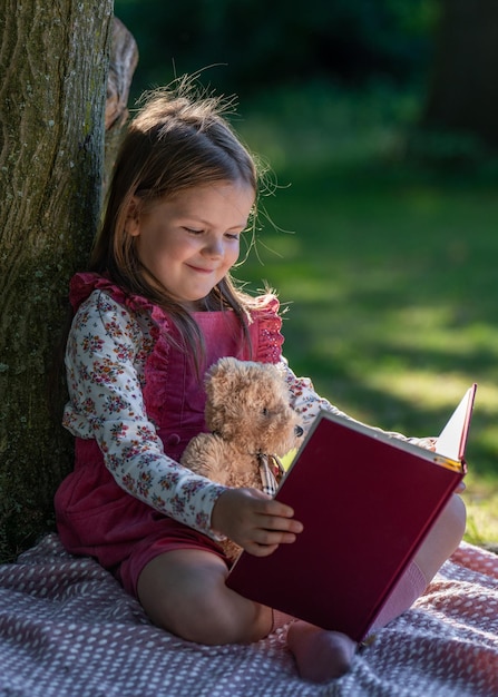 Una niña pequeña se sienta cerca de un gran árbol en el parque y lee un libro, un pequeño osito de peluche se sienta a su lado