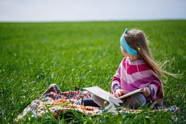 La niña pequeña se sienta en una colcha y lee un libro con un cuento de hadas, hierba verde en el campo, clima primaveral soleado, sonrisa y alegría del niño, cielo azul con nubes