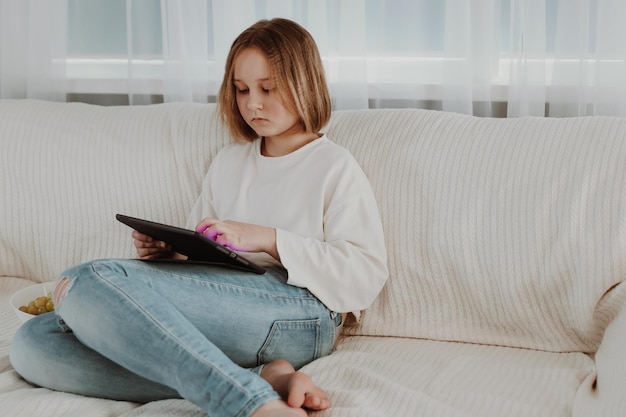 La niña pequeña se sienta en un cómodo sofá disfrutando de jugar en línea en una tableta digital Adicto a la tecnología