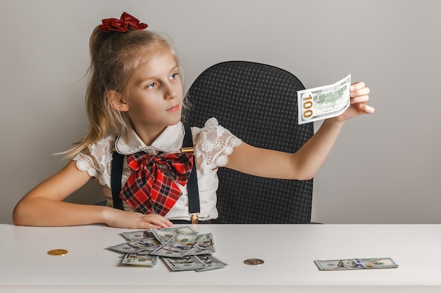 Una niña pequeña se sienta en una mesa frente a una pila de billetes de cien dólares y examina uno de ellos en busca de controles ligeros de falsedad.