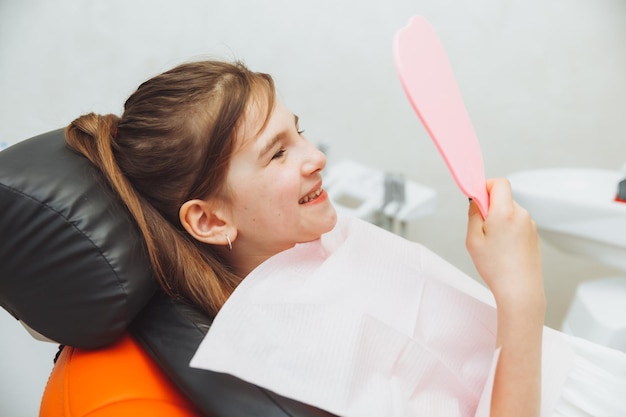 Una niña pequeña se sienta en una silla dental y examina sus dientes curados mientras se mira en el espejo