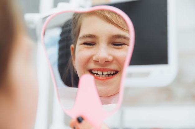 Una niña pequeña se sienta en una silla dental y examina sus dientes curados mientras se mira en el espejo