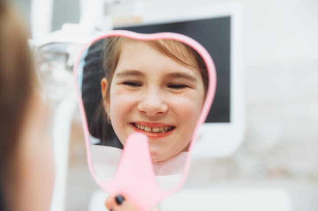 Una niña pequeña se sienta en una silla dental y examina sus dientes curados mientras se mira en el espejo