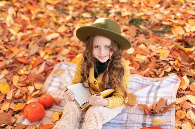 Una niña pequeña se sienta en una tela escocesa con calabazas y lee un libro sobre un fondo otoñal