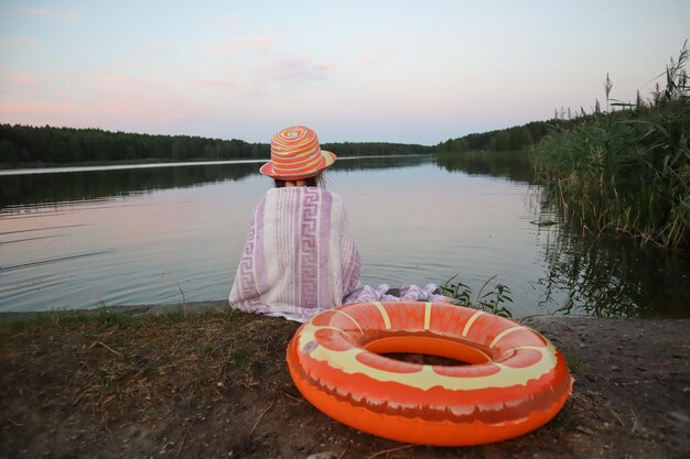 Una niña pequeña con un sombrero amarillo cubierta con una toalla se sienta en la orilla del lago junto a un círculo inflable por la noche de verano, el concepto de unas hermosas vacaciones de verano para los niños