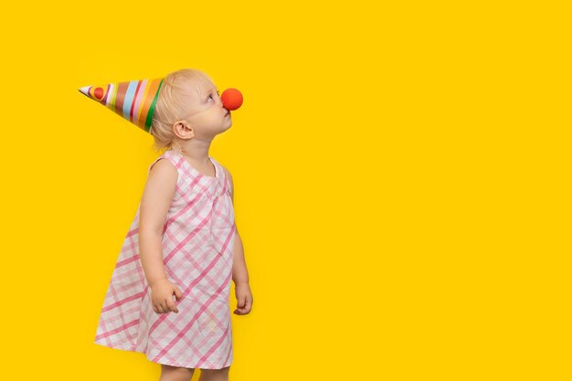 Niña pequeña con sombrero de fiesta y nariz de payaso roja y mira hacia arriba Retrato sobre fondo amarillo Copiar espacio