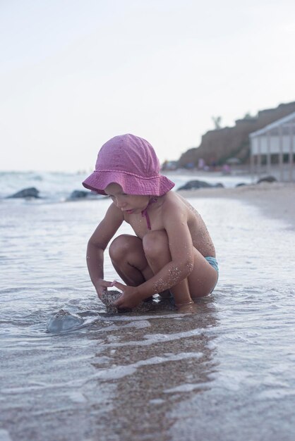 La niña pequeña con un sombrero se juega en el agua en la playa.