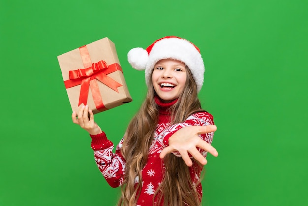 Una niña pequeña con un sombrero y un suéter de Santa Claus estira la mano hacia adelante y sostiene un regalo de Navidad en un fondo verde aislado Un regalo de Año Nuevo para un niño