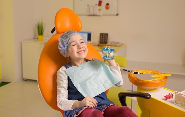 Una niña pequeña sonríe y sostiene un modelo de mandíbula en sus manos mientras se sienta en una silla en una clínica dental Cuidado dental higiene bucal