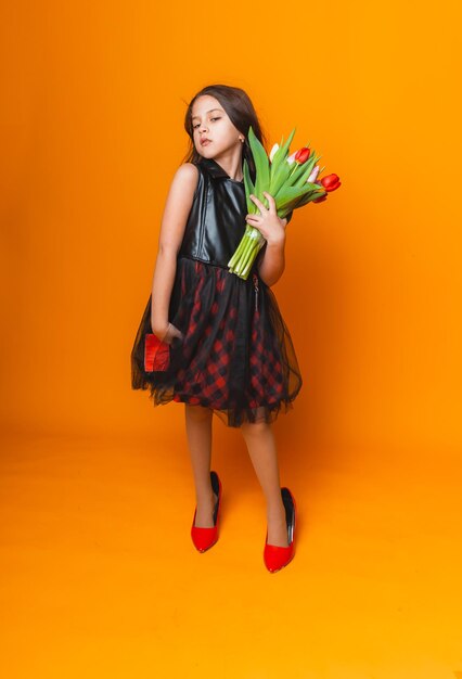 Niña pequeña sonríe con un vestido y grandes zapatos rojos sostiene un ramo de flores en un espacio de fondo amarillo para texto