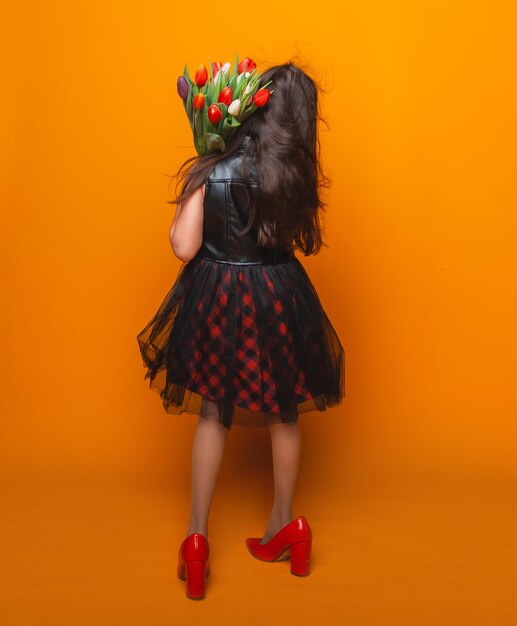 Niña pequeña sonríe con un vestido y grandes zapatos rojos sostiene un ramo de flores en un espacio de fondo amarillo para texto