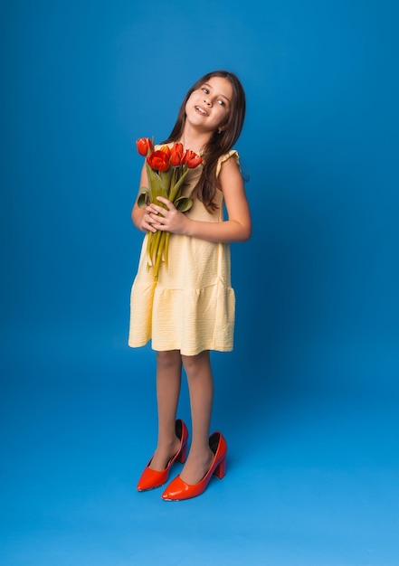 Niña pequeña sonriendo con un vestido y grandes zapatos rojos sosteniendo un ramo de flores en un espacio de fondo azul para el texto
