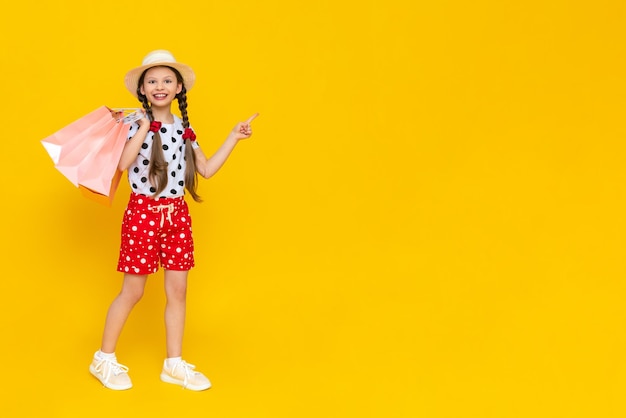 Una niña pequeña sostiene bolsas de compras Un niño con un sombrero de verano y pantalones cortos se dedica a las compras de los niños Un niño feliz en pleno crecimiento sobre un fondo amarillo aislado Banner Copiar espacio