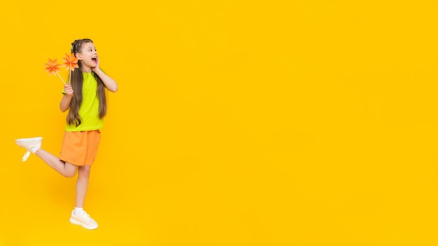 Una niña pequeña sostiene flores de origami en un palo y se mantiene erguida La creatividad de los niños Un niño en pantalones cortos y una camiseta sueña con un verano soleado en un fondo amarillo aislado Banner Copiar espacio