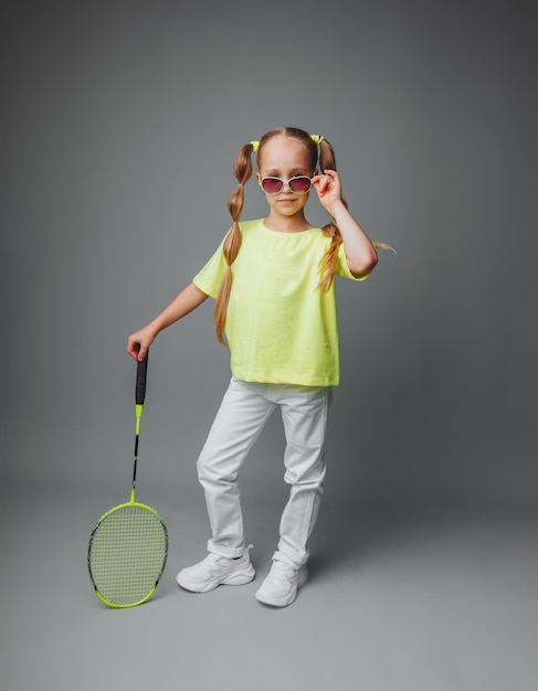 Una niña pequeña sostiene una raqueta sobre un fondo gris, un pequeño jugador de tenis, un niño pequeño está jugando al tenis, deportes infantiles.
