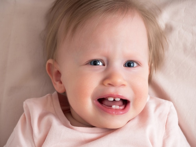 Una niña pequeña sufre de dolor, dentición, mirando a la cámara, llorando, primer plano. Retrato de una hermosa niña de luz en tonos rosados. Niño de un año de edad.