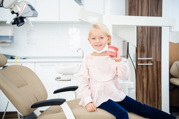 Una niña pequeña tiene dentaduras postizas artificiales en la mano en el consultorio dental