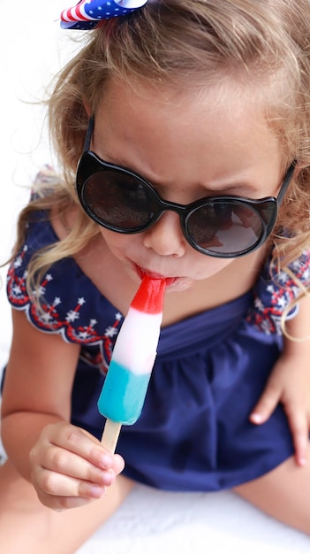 Niña pequeña con traje de baño azul comiendo un helado en colores rojo blanco y azul