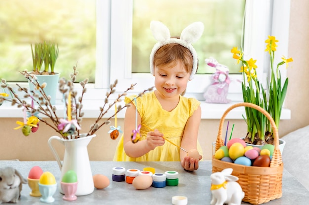 Una niña pequeña vestida de amarillo con orejas de conejo en la cabeza y pintando huevos de Pascua de buen humor