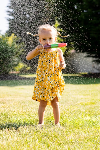 Una niña pequeña con un vestido amarillo con flores en la cabeza está rociando agua en su aspersor.